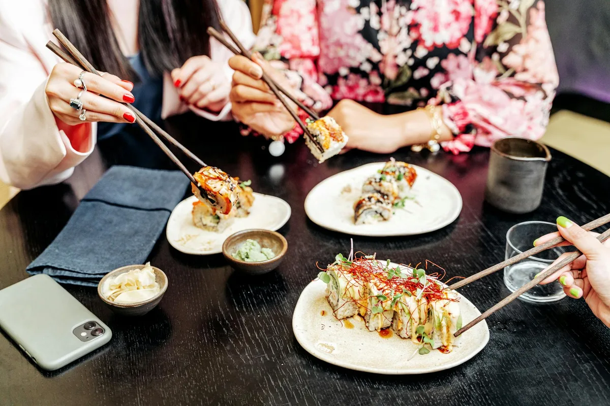 A group of people dining together, enjoying sushi with chopsticks and various condiments on a table.
