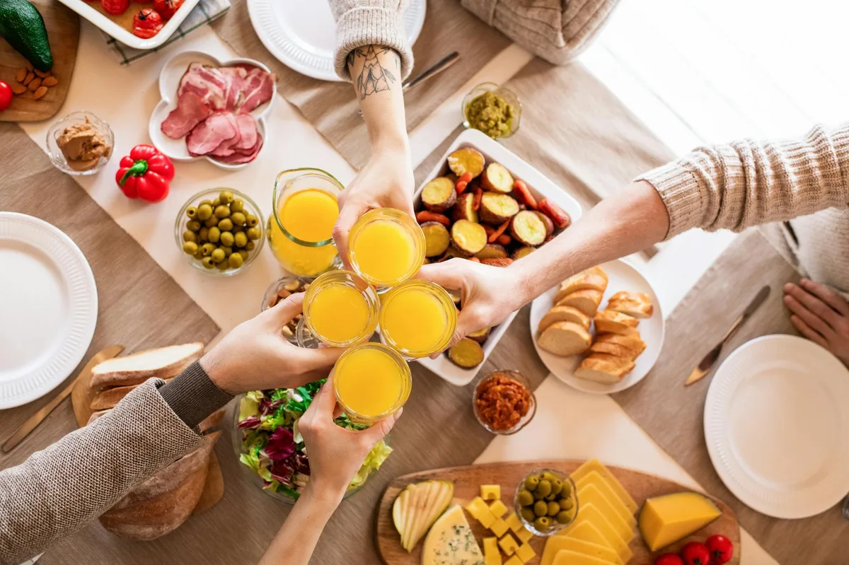 Top view of friends toasting with orange juice during a brunch with diverse foods.